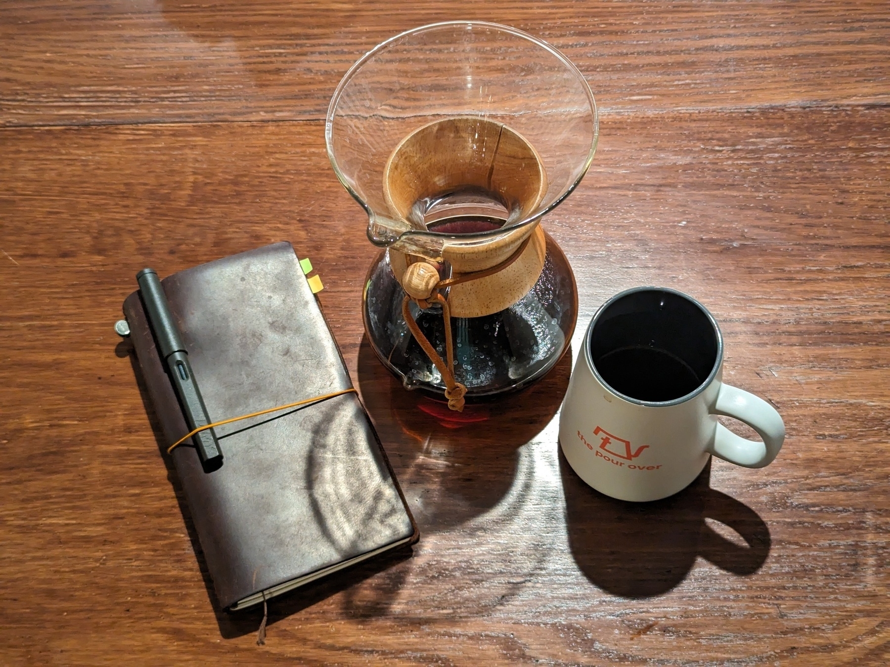 A brown leather journal with a black pen clipped on, a Chemex pour-over and a coffee mug sit atop a brown wooden table.