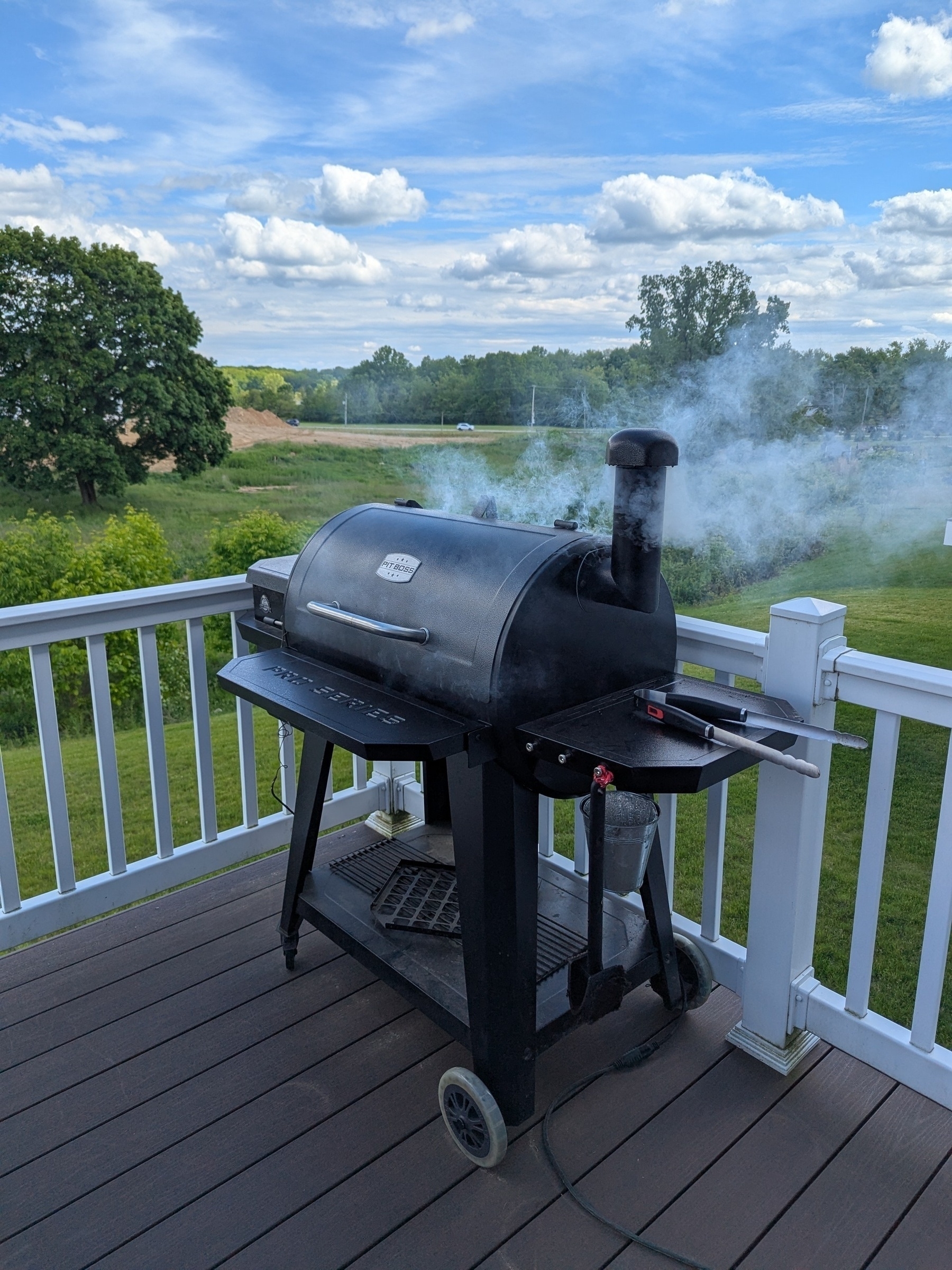 A smoker grill is actively smoking on a wooden deck overlooking a green field and trees under a cloudy sky.